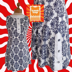 May include: A vintage sleeveless dress with a white collar and button detail. The dress has a blue and white floral pattern. The Tank Top Vintage logo is visible. The dress is displayed on a mannequin.