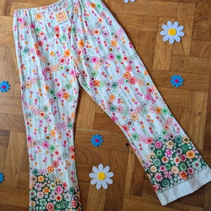 May include: White floral print pants with a colorful flower pattern. The pants have a flared leg and an elastic waistband. The bottom of the pants have a green floral border. The pants are laying on a wooden floor with flower decorations.
