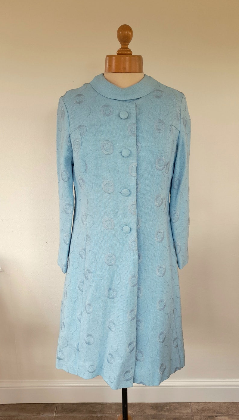 May include: A light blue dress with a round, embroidered pattern and button fastening. The dress has long sleeves and a fitted silhouette.