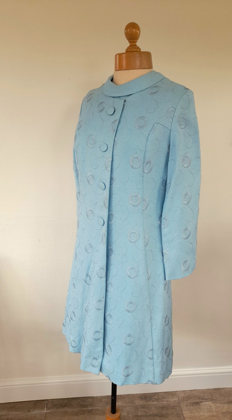 May include: A light blue dress with a round, embroidered pattern. The dress has a collar and a button-up front.