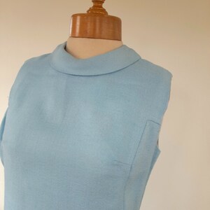 May include: A light blue sleeveless dress with a mock neck collar. The dress is on a wooden dress form.