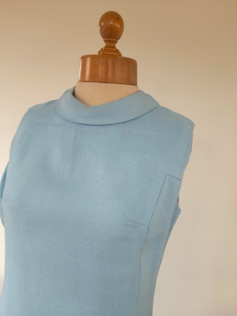 May include: A light blue sleeveless dress with a mock neck collar. The dress is on a wooden dress form.