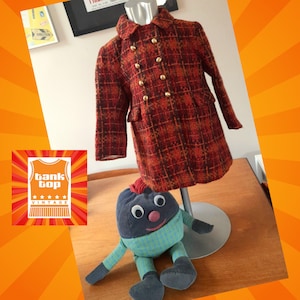 May include: A red and black checked coat with gold buttons. The coat is on a mannequin and there is a small blue and green toy in the foreground.
