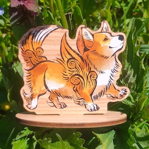 Peut inclure: Figurine en bois de corgi avec fourrure orange et blanche, ornée d'ailes décoratives. Le corgi est posé sur un socle rond en bois. La figurine est placée sur un fond d'herbe verte et de feuillage.