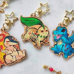 Puede incluir: Llaveros de madera con ilustraciones coloridas de tres criaturas de dibujos animados. Cada llavero tiene un cierre dorado en forma de estrella. Las criaturas son en tonos de rojo, amarillo, azul y verde.