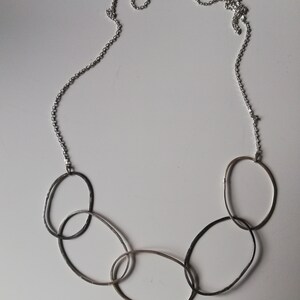 May include: Silver-toned necklace featuring a series of interconnected oval-shaped links. The necklace has a delicate chain and a cluster of textured, elongated ovals. The design is simple and modern, suitable for everyday wear.