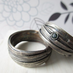 Op de afbeelding: Twee zilveren ringen met een donkere, geoxideerde afwerking en een getextureerd, gelaagd oppervlak. Eén ring is voorzien van een kleine, ronde, donkergroene edelsteen in de zetting. De banden hebben een ruw uiterlijk met zichtbare horizontale groeven.