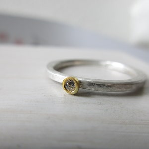 Könnte beinhalten: Ein silberner Ring mit einer strukturierten Oberfläche und einem kleinen, runden, klaren Edelstein, der in einer goldenen Fassung sitzt.