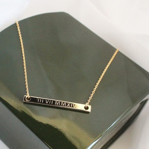 May include: A gold chain necklace with a thin bar pendant engraved with the Roman numerals "III VII MMXIV".