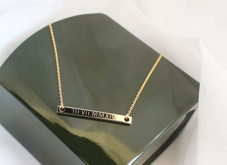 May include: A gold chain necklace with a thin bar pendant engraved with the Roman numerals "III VII MMXIV".