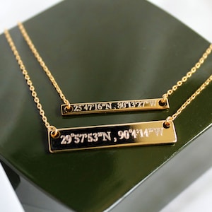 May include: Two gold necklaces with engraved bar pendants. The top pendant reads "25°47'16"N, 80°13'27"W" and the bottom pendant reads "29°57'53"N, 90°4'14"W".