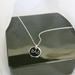 May include: A silver chain necklace with a black round pendant that says "ΦΑΔ" in white lettering.