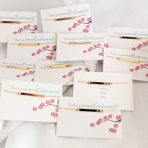 May include: A set of gold bar bracelets with the text "Thank you for being my Bridesmaid!" on white cards with pink cherry blossom designs.