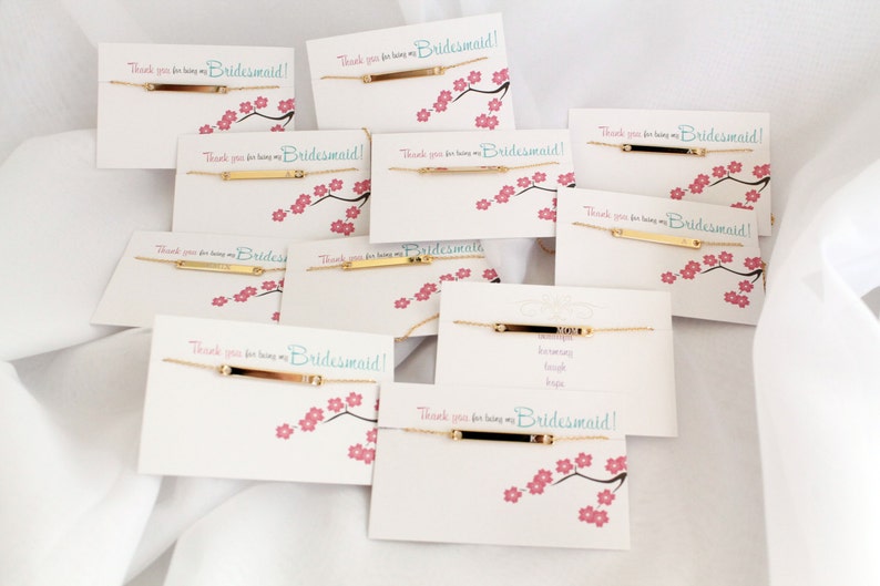 May include: A set of gold bar bracelets with the text "Thank you for being my Bridesmaid!" on white cards with pink cherry blossom designs.