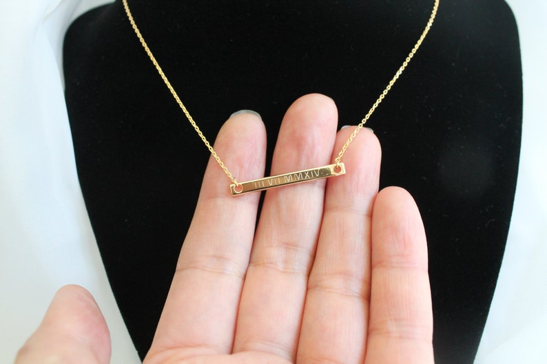 May include: A gold chain necklace with a thin gold bar pendant engraved with the Roman numerals "II VII MMXIV".