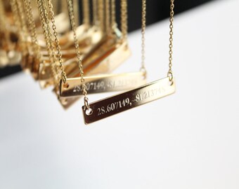 Op de afbeelding: Een gouden ketting met een rechthoekige hanger gegraveerd met de coördinaten "28.607149, -81.213748".