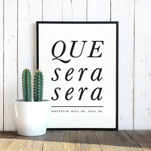 Könnte beinhalten: Ein schwarz-weiß gerahmter Druck mit den Worten "QUE sera sera" in einer stilvollen Schriftart. Darunter steht der Satz "WHATEVER WILL BE, WILL BE." Links vom Druck steht ein kleiner weißer Topf mit zwei Kakteen.