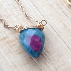 May include: A gold chain necklace with a teardrop-shaped blue gemstone pendant with a pink center. The pendant is wrapped in gold wire.