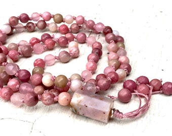 May include: A pink gemstone mala necklace with a large pink gemstone pendant and a tassel. The necklace is made of small, round beads and is strung on a pink cord.
