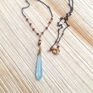 Op de afbeelding: Een delicate ketting met een traanvormige blauwe edelsteen hanger. De ketting heeft een zilveren ketting met kleine rode kralen en een gouden draadwikkeling rond de hanger.