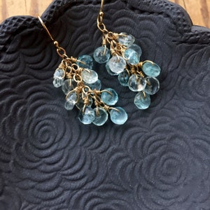 May include: A pair of gold dangle earrings with light blue gemstone drops. The earrings are hanging from a black surface with a floral pattern.
