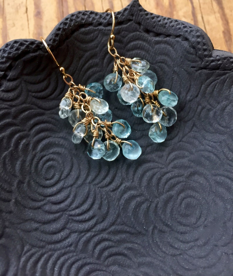 May include: A pair of gold dangle earrings with light blue gemstone drops. The earrings are hanging from a black surface with a floral pattern.