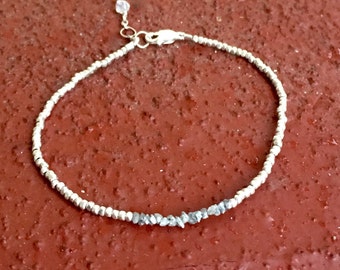 Puede incluir: Una pulsera de plata con una fila de pequeños diamantes grises en bruto. La pulsera está hecha de pequeñas cuentas de plata y tiene un cierre de plata.