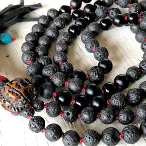 Może przedstawiać: Czarny naszyjnik mala z lawy wulkanicznej i czarnego onyksu ze sznurkiem czerwonym i brązową drewnianą koralikiem na końcu. Naszyjnik leży na białym drewnianym tle.