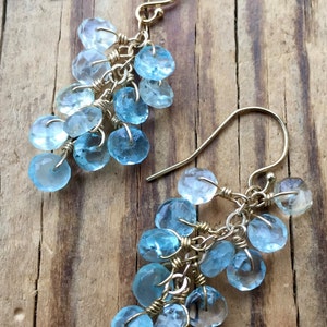 May include: A pair of gold dangle earrings with light blue teardrop-shaped gemstones. The earrings are made with wire wrapped around the gemstones, creating a cascading effect.