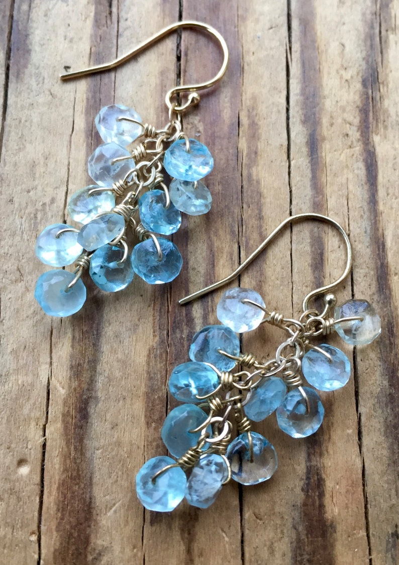 May include: A pair of gold dangle earrings with light blue teardrop-shaped gemstones. The earrings are made with wire wrapped around the gemstones, creating a cascading effect.