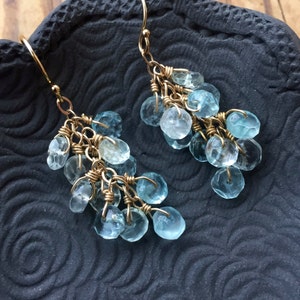 May include: A pair of gold dangle earrings with light blue gemstone drops. The earrings are made with multiple small, faceted gemstones that are connected by gold wire.
