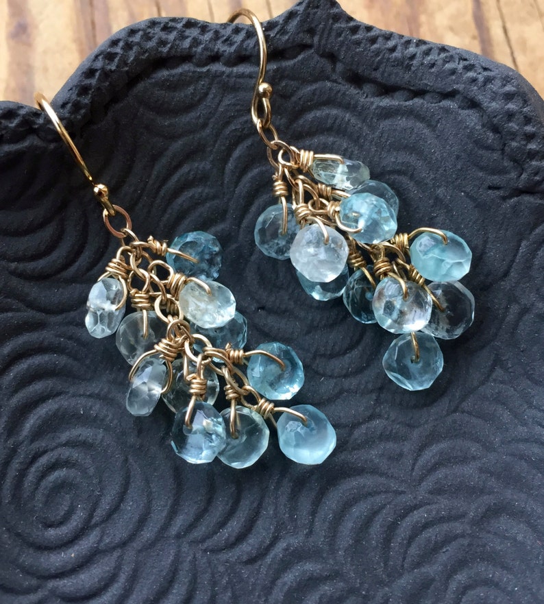 May include: A pair of gold dangle earrings with light blue gemstone drops. The earrings are made with multiple small, faceted gemstones that are connected by gold wire.