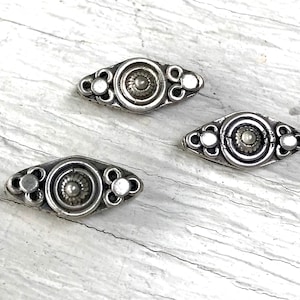 May include: Three silver metal decorative embellishments with a circular design in the center and three smaller circles on each side. The embellishments are in the shape of an oval.