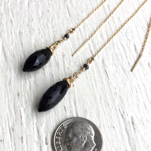 May include: A pair of black gemstone dangle earrings with gold chains and small black beads. The earrings are shown next to a US dime for scale.