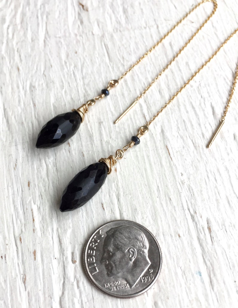 May include: A pair of black gemstone dangle earrings with gold chains and small black beads. The earrings are shown next to a US dime for scale.