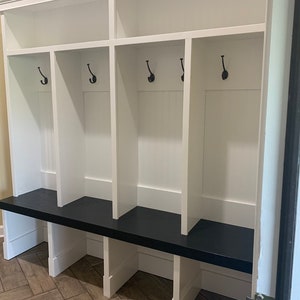 May include: White wooden storage bench with a black top and four cubbies. Each cubby has a hook and a shelf. The bench is in a hallway with a tiled floor.