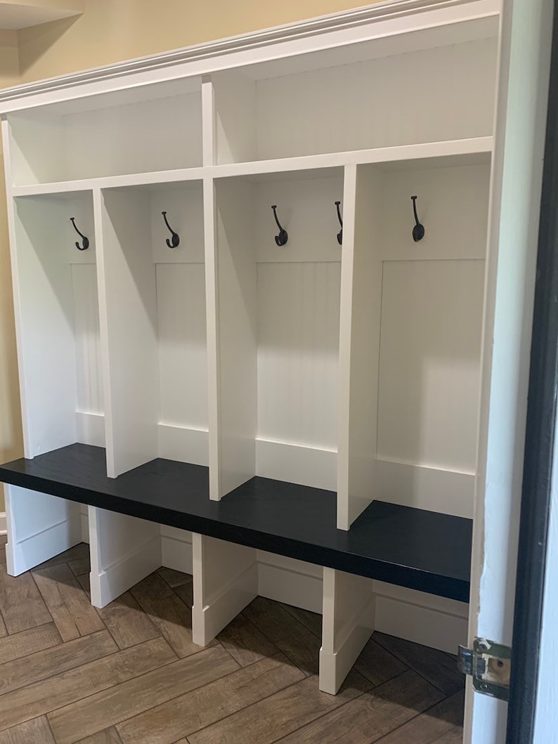 May include: White wooden storage bench with a black top and four cubbies. Each cubby has a hook and a shelf. The bench is in a hallway with a tiled floor.