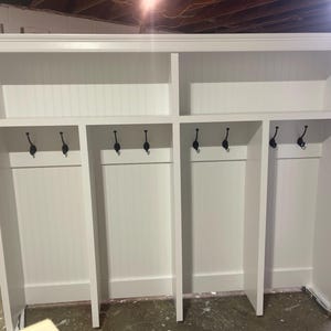 May include: White wooden entryway storage unit with four cubbies and black hooks. The unit has a shelf above each cubby.