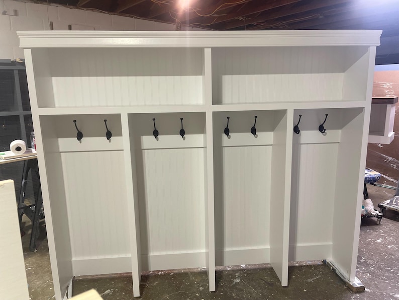 May include: White wooden entryway storage unit with four cubbies and black hooks. The unit has a shelf above each cubby.