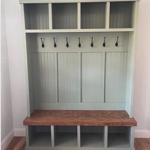 May include: A sage green entryway storage unit with a wooden bench and cubby storage. The unit has six black hooks, four upper shelves, and four lower cubbies. The bench is made of stained wood.