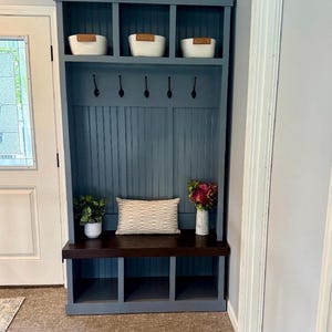 Può includere: Unità di stoccaggio blu per l'ingresso con panca, ganci e ripiani. Tre cesti bianchi sono sullo scaffale superiore. Un cuscino decorativo, una pianta e un vaso con fiori sono sulla panca. L'unità è contro un muro grigio.