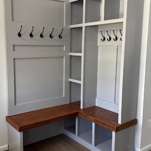 May include: A custom-built, gray corner entryway bench with a dark wood seat and back. The unit features multiple storage cubbies, coat hooks, and open shelving. The design is modern and functional, perfect for organizing a home's entrance.