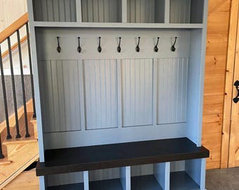 May include: A gray and dark brown wooden entryway bench with storage cubbies, a bench seat, and a row of hooks. The unit has a top shelf with multiple compartments and vertical paneling on the back.
