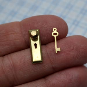 以下が含まれることがあります： 金色のミニチュアのドアノブと鍵。ノブは丸いハンドルと鍵穴があります。鍵は丸い頭を持つシンプルなデザインです。背景には「navy blue navel miniature handles & door knobs」というテキストが表示されています。