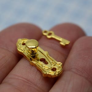 以下が含まれることがあります： 金色調のミニチュアのドアノブとキーのセット。ノブは装飾的なデザインと丸いハンドルが付いています。キーはシンプルで、上部に小さなループが付いています。