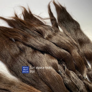 Könnte beinhalten: Ein Bündel brauner Suri-Alpaka-Locken, die mit schwarzen Gummibändern zusammengebunden sind. Der Text "Suri alpaca locks PER" ist auf dem Bild sichtbar.
