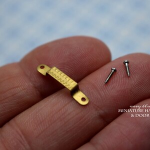 May include: A gold-colored miniature door handle with a decorative pattern. The handle is held in a hand, along with two small silver screws. The text "navy blue navel MINIATURE HANDLES & DOOR KNOBS" is visible below the handle.