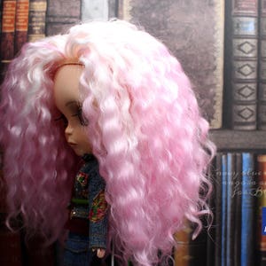 May include: A doll with long, curly pink and white hair. The doll is wearing a denim jacket and jeans. The doll is standing in front of a bookcase.