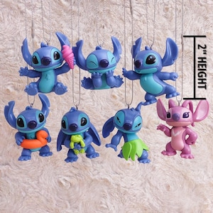May include: A collection of blue and pink character ornaments, each approximately 2 inches in height, hanging from silver chains. The ornaments depict a popular cartoon character in various poses, some holding accessories like an ice cream cone or a frog.