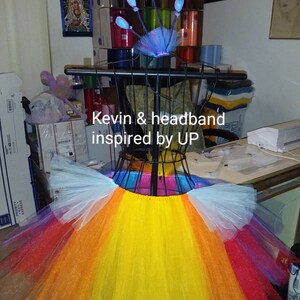 May include: A multi-colored tulle tutu skirt with a rainbow pattern. The skirt is on a mannequin and has a white, yellow, orange, red, and purple layers. The text "Kevin & headband inspired by UP" is visible in the image.